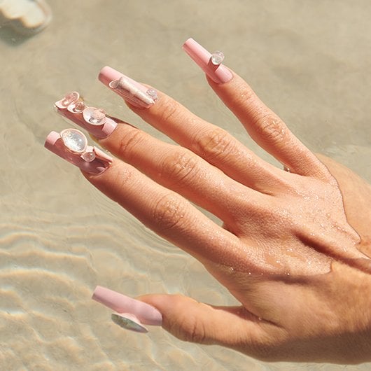 Model hand with a light pink manicure featuring 3D nail art against a sandy beach background