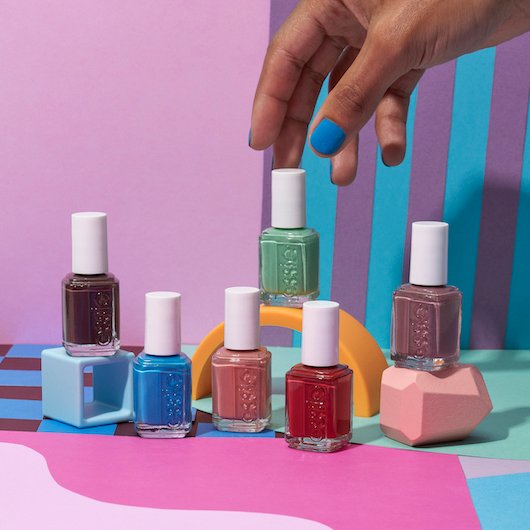 Dark skin model's hand reaching for a bottle of green essie nail polish surrounded by other bottles